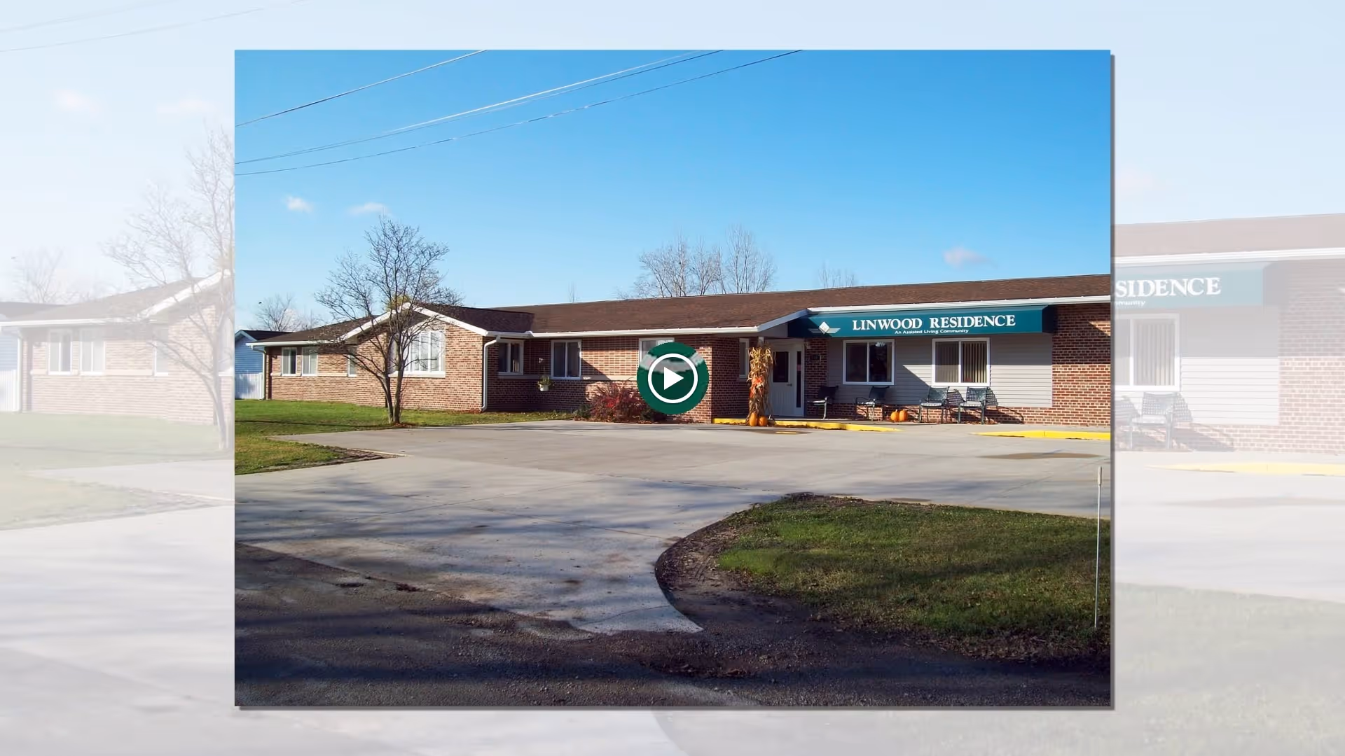 Exterior view of Linwood Residence, a single-story brick assisted living facility with a paved driveway and a small grassy area in front. The building has a green awning with the text 'LINWOOD RESIDENCE' above the entrance. There are a few chairs and some autumn decorations near the entrance.