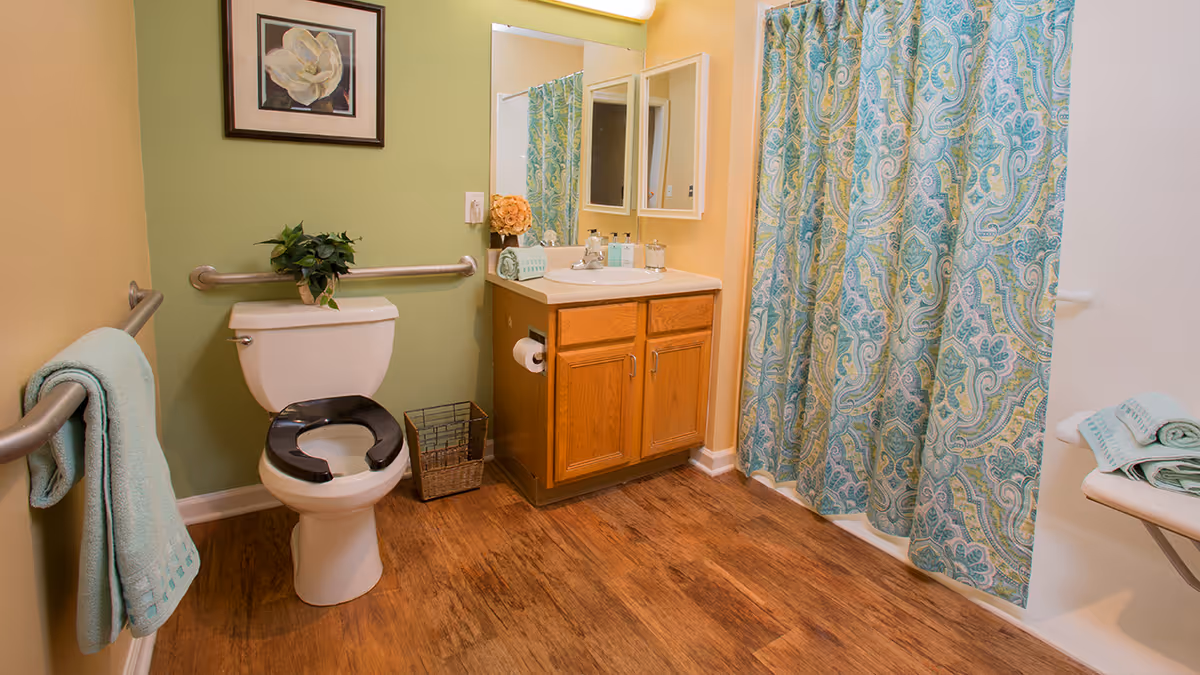 A bathroom with a white toilet featuring a black raised toilet seat, a wooden vanity with a sink and mirror, a patterned blue and green shower curtain, wooden floor, and light green and yellow walls. There are handrails on the walls and a towel hanging on one of them. A framed flower picture hangs above the toilet, and a small plant is placed on the toilet tank.