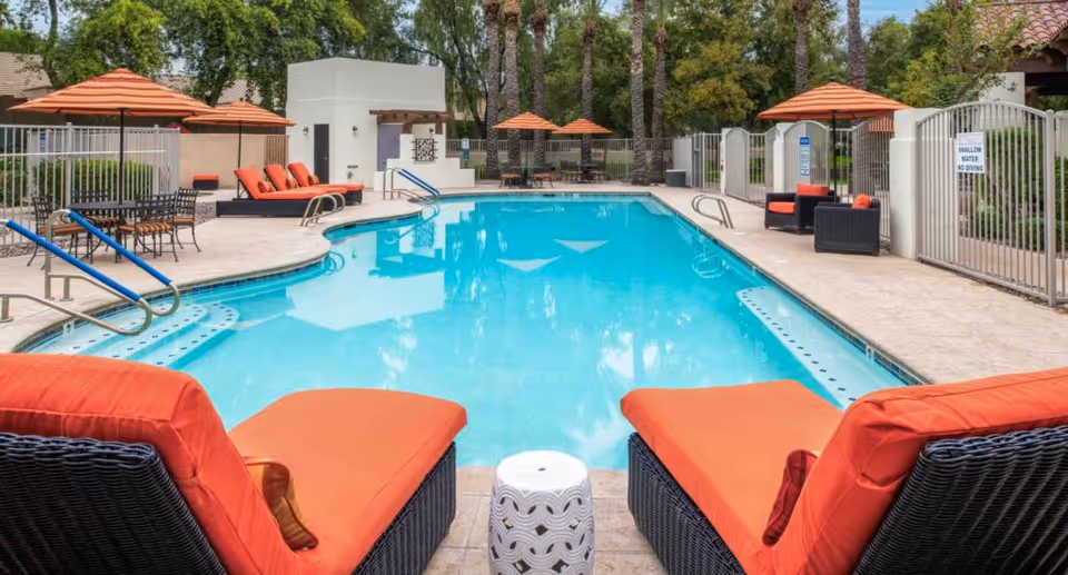 Outdoor swimming pool area with clear blue water, surrounded by lounge chairs with orange cushions and matching orange umbrellas. There are tables and chairs on the left side, and a gated fence enclosing the pool area with trees and greenery in the background.