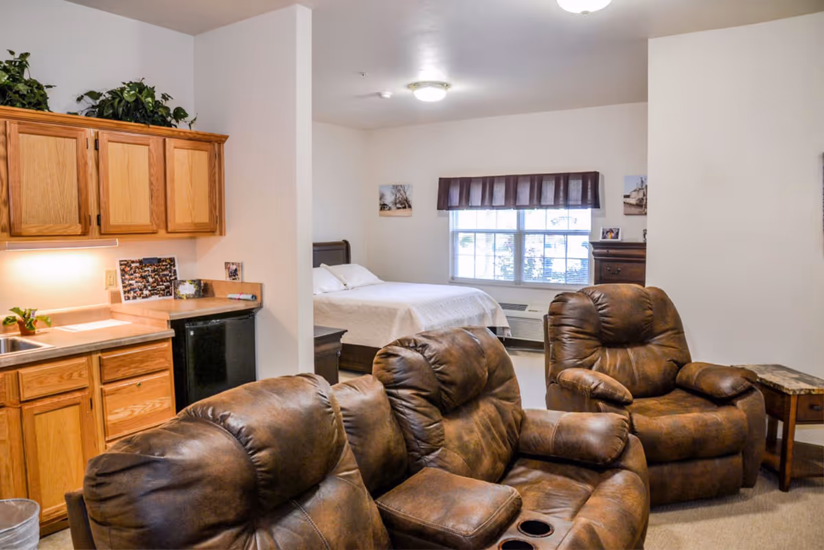Interior view of a senior living facility room featuring a small kitchenette with wooden cabinets and a sink on the left, a cozy seating area with brown leather recliners in the foreground, and a bedroom area with a bed, window with blinds and valance, and a dresser in the background.