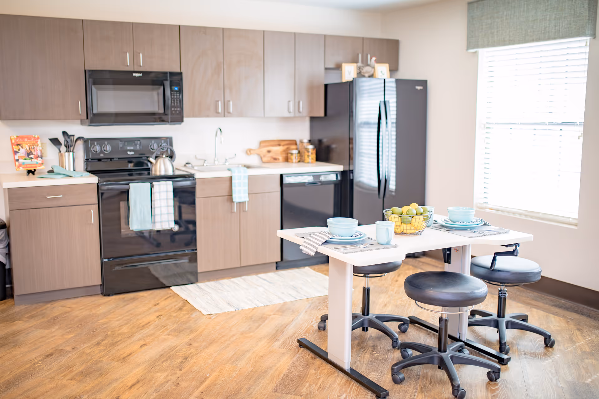 Bright modern kitchen with wood cabinets, black stove and refrigerator, a small dining table set with dishes and four rolling stools.