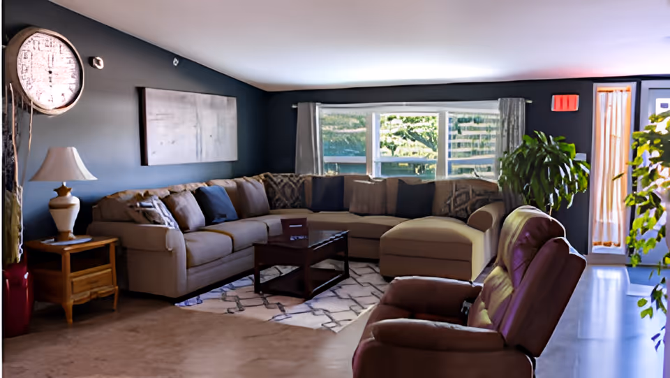 A cozy living room with a large beige sectional sofa adorned with multiple cushions, a wooden coffee table on a patterned rug, a brown recliner chair in the foreground, a wooden side table with a lamp, a large wall clock, and a window with curtains letting in natural light. There are also green plants near the window and door.
