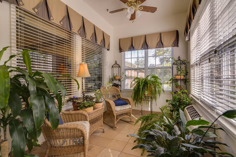 A bright sunroom with large windows covered by blinds and valances. The room contains wicker furniture including two chairs and a small table, several potted plants, a floor lamp, and a ceiling fan with wooden blades. The floor is tiled and sunlight streams through the windows.