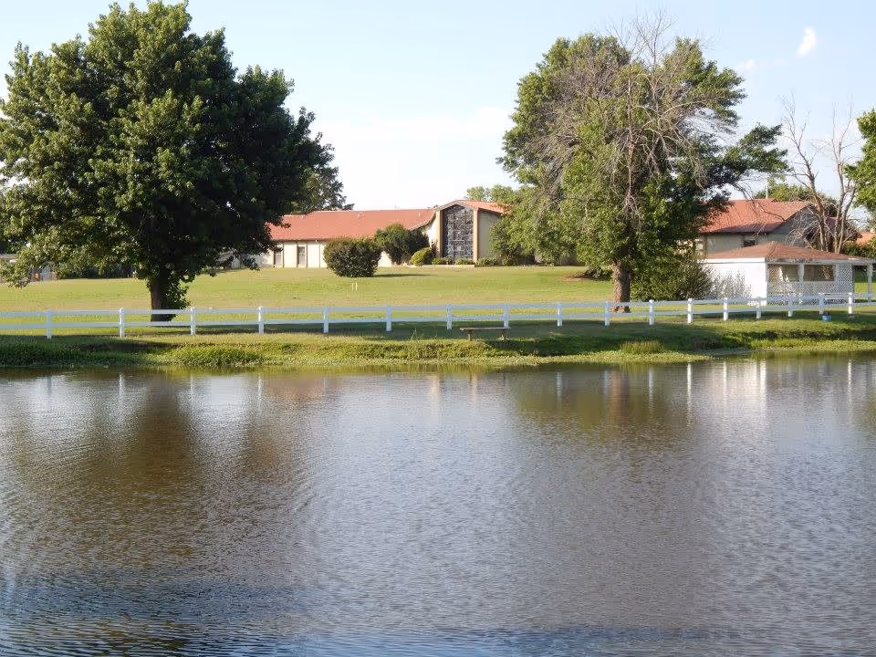 A serene outdoor scene at Franciscan Villa featuring a calm pond in the foreground, a white fence, green grass, and several large trees. In the background, there is a single-story building with a red roof partially obscured by trees.