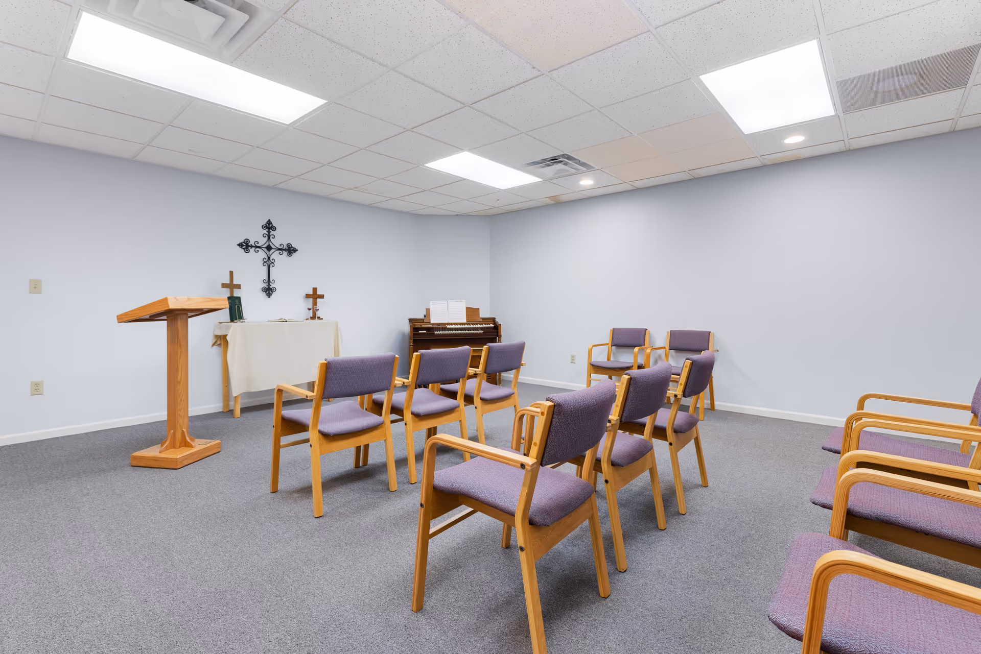 A small chapel or prayer room with a wooden lectern, a table covered with a white cloth holding two crosses and a book, a piano with sheet music, and several wooden chairs with purple cushions arranged in rows on a gray carpeted floor under a white tiled ceiling with fluorescent lights.