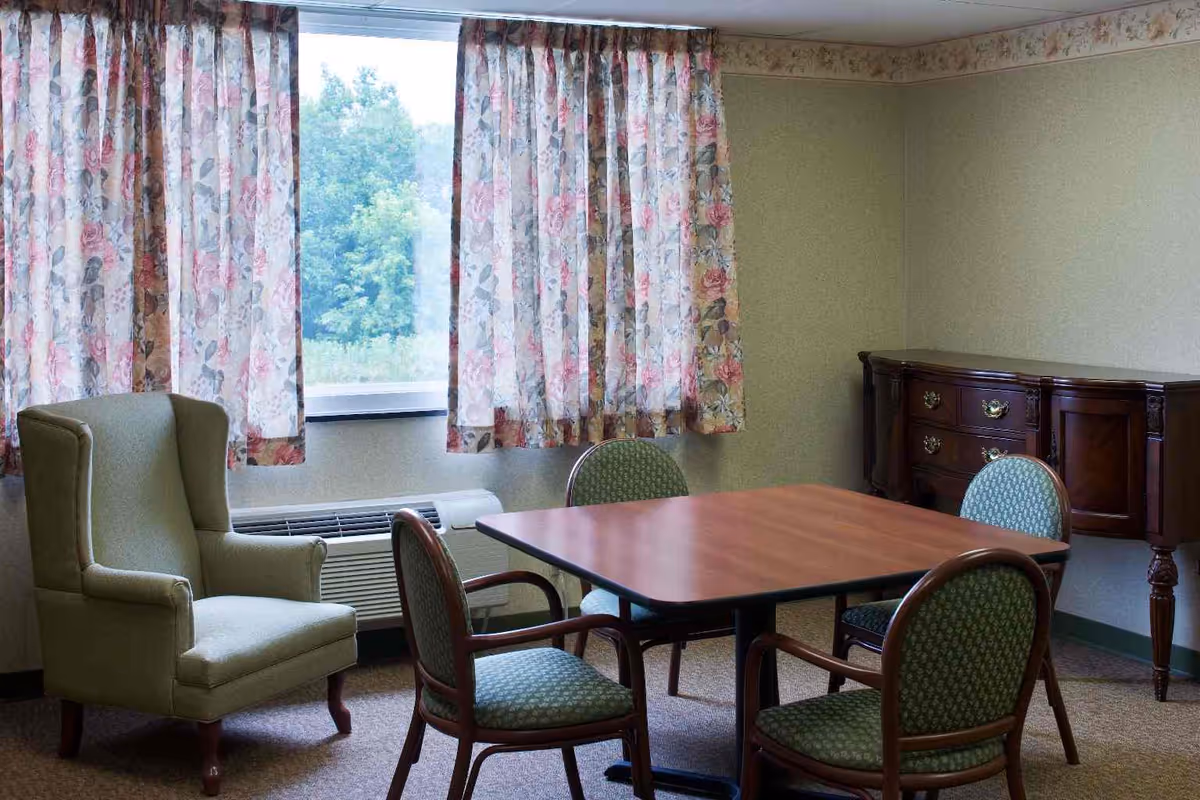 A small sitting area with a square wooden table surrounded by four green upholstered chairs with wooden arms. To the left is a green upholstered wingback armchair. Behind the table is a window with floral patterned curtains, and beneath the window is an air conditioning unit. On the right side of the room is a dark wooden sideboard with drawers and decorative handles. The walls are light-colored with a floral border near the ceiling.