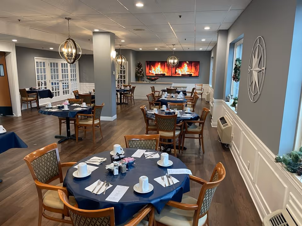 A dining room with multiple round tables covered in dark blue tablecloths, each set with white cups, saucers, napkins, and silverware. The room has wooden chairs with patterned cushions, wood flooring, and gray walls. There are modern spherical pendant lights hanging from the ceiling and a large digital fireplace display on the far wall. Windows on the right side have holiday wreath decorations.