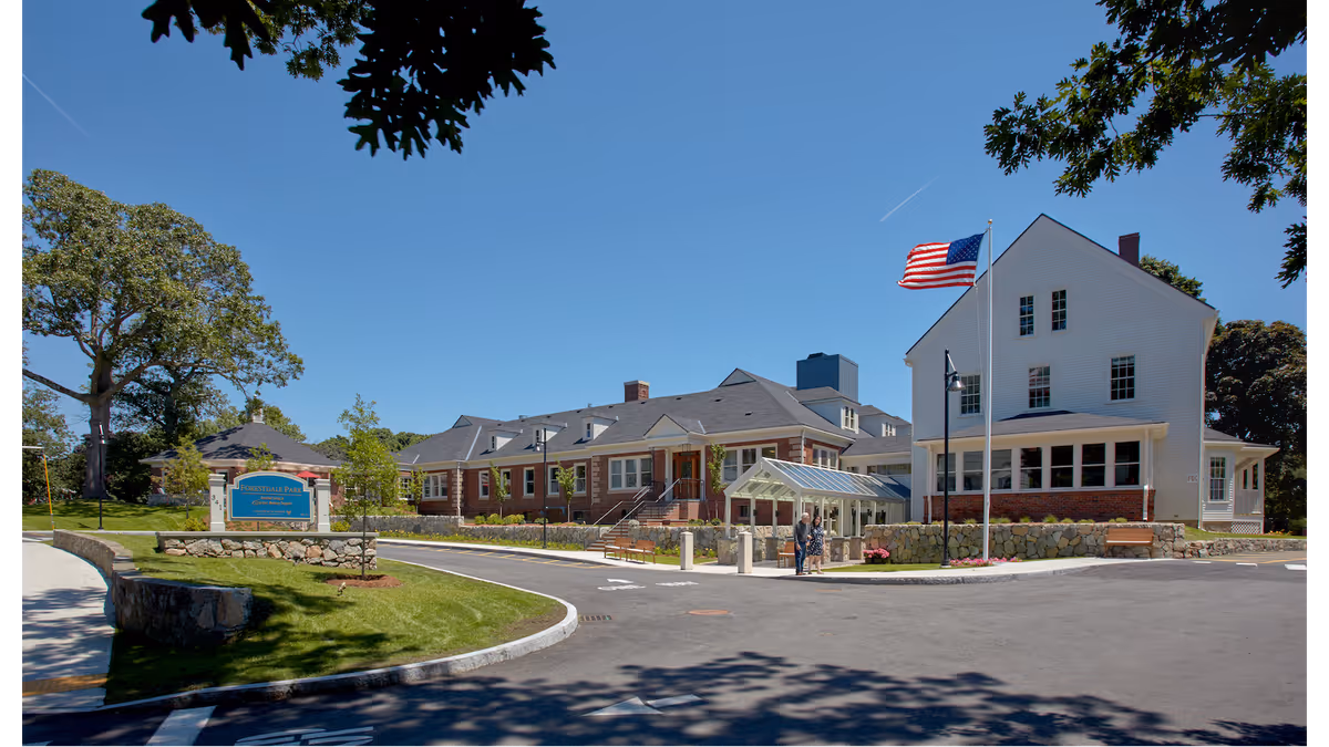 Exterior view of Forestdale Park Senior Living facility on a clear sunny day, showing a large building with white and brick walls, an American flag on a flagpole, a curved driveway, green lawn, trees, and a sign for Forestdale Park Senior Living near the entrance.