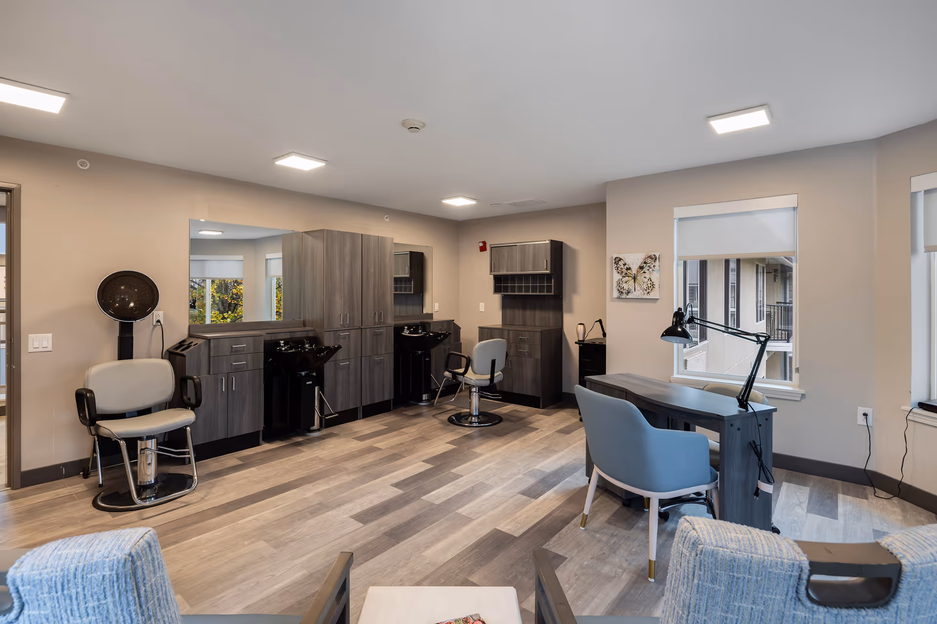 A modern salon area inside a senior living facility featuring two hair washing stations with chairs, a hair dryer chair, a manicure table with a lamp and chair, and large windows letting in natural light. The room has wood flooring and neutral-colored walls.