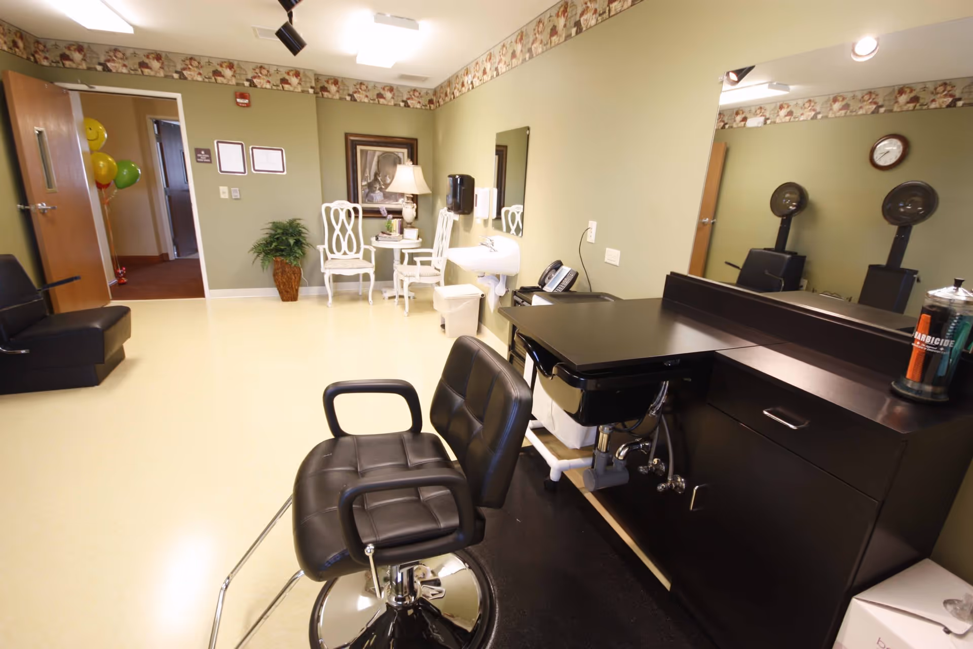 Interior view of a salon area in a senior living facility with a black salon chair in front of a black counter and sink. The room has light green walls with a decorative border near the ceiling, a large mirror, a small white table with two white chairs, a lamp, a plant, and a framed picture on the wall. There are balloons visible in an adjacent room through an open door.