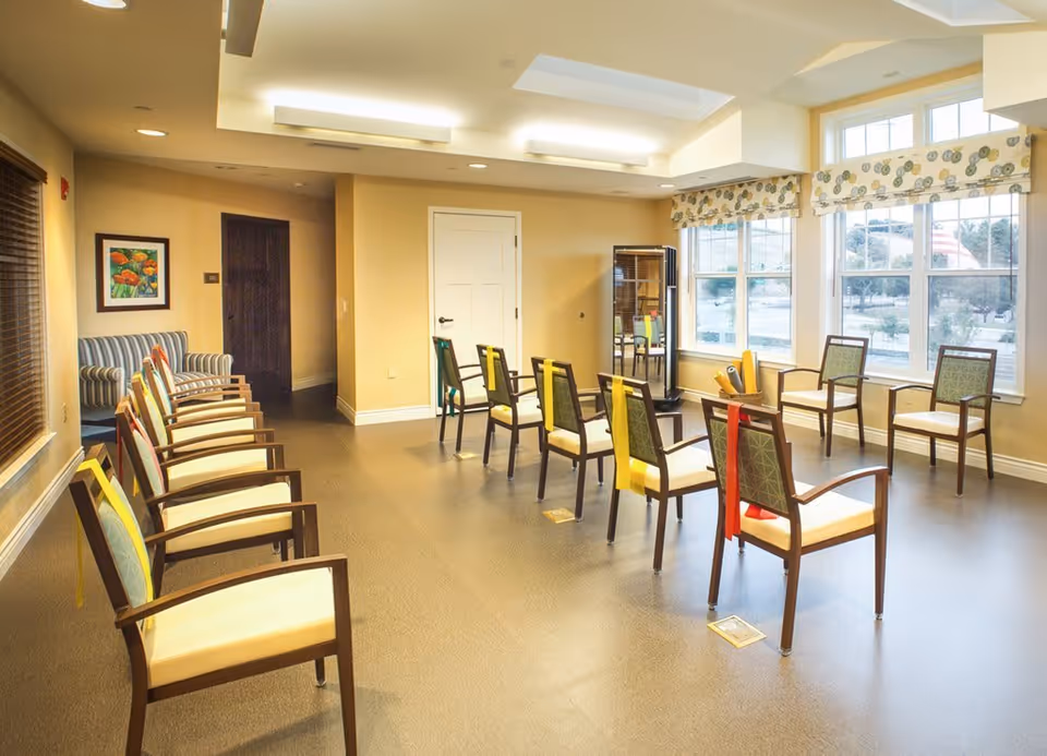 A well-lit room with multiple chairs arranged in a spaced-out manner, some with colored resistance bands draped over the backs. The room has large windows with patterned valances, a striped armchair in the corner, and a framed floral painting on the wall. The floor is smooth and the walls are painted a soft beige color.
