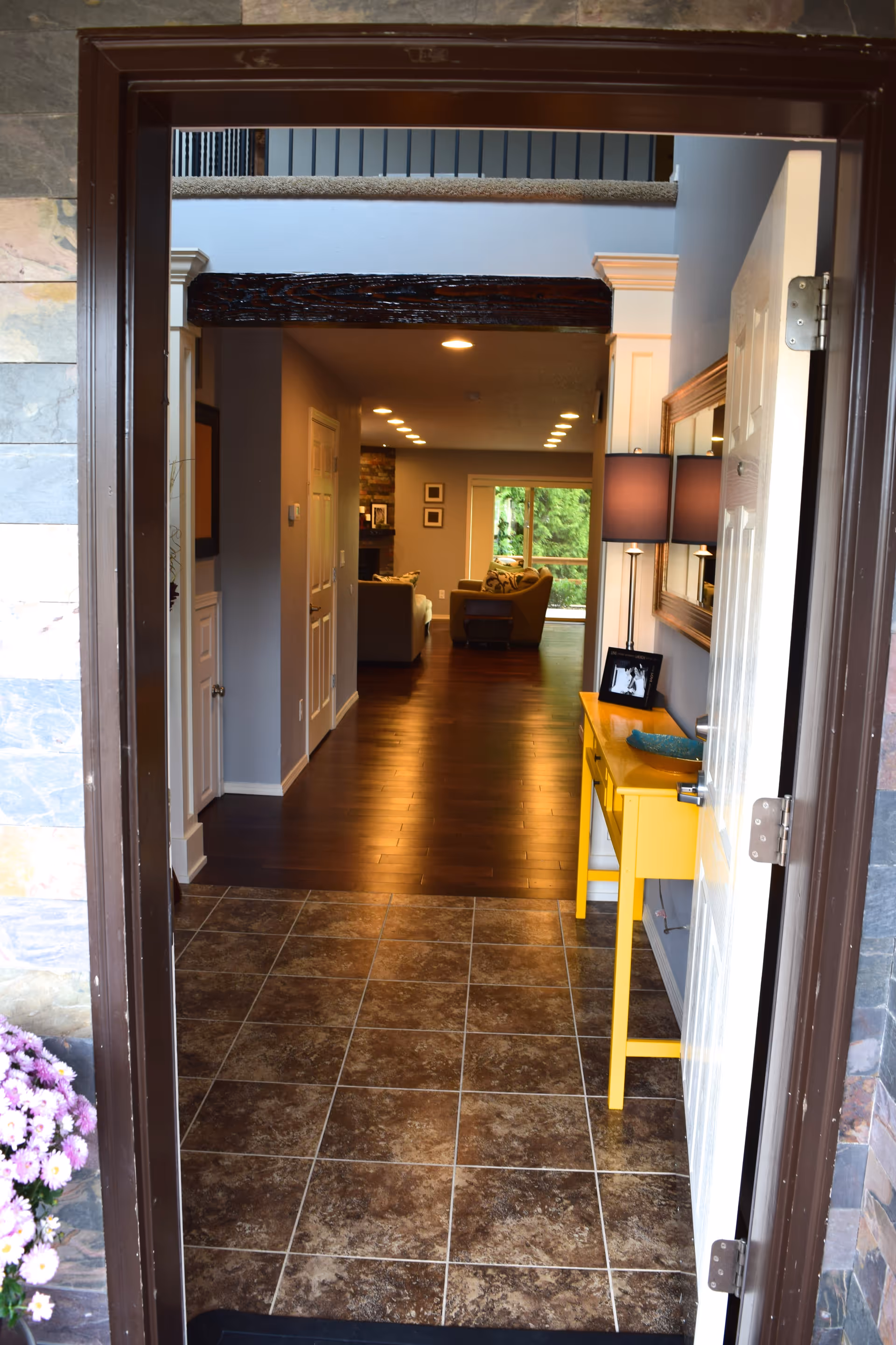 View through an open front door into a home interior showing a tiled entryway leading to a hallway with wooden flooring. A yellow console table with a lamp and framed photo is on the right side. The hallway leads to a living room area with sofas and a large window overlooking greenery.