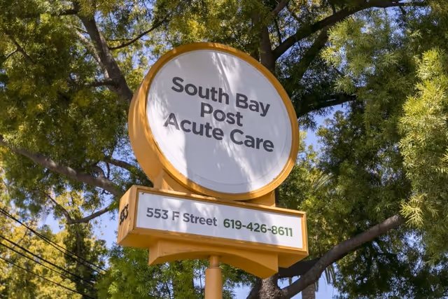 Round yellow sign reading "South Bay Post Acute Care" with the address and phone number on a post in front of leafy trees.