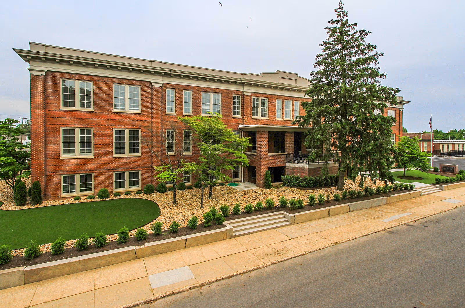 Exterior view of a three-story brick building with multiple windows, surrounded by landscaped greenery including small trees, bushes, and a large pine tree. The building has a small covered entrance and is adjacent to a sidewalk and street.