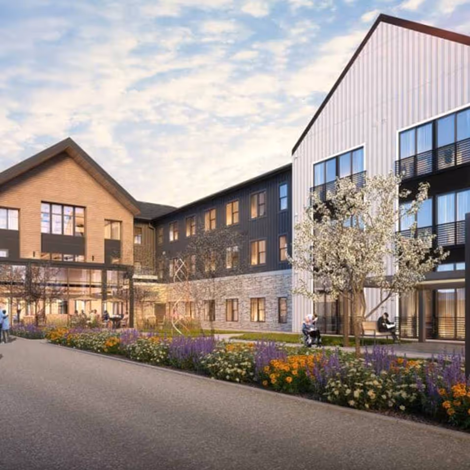 Exterior view of a senior living facility with a paved walkway, landscaped flower beds with purple, orange, and white flowers, and a few trees. The building has multiple stories with large windows, featuring a mix of wood, stone, and metal siding. Several people are visible outside, including one person in a wheelchair and another sitting on a bench.