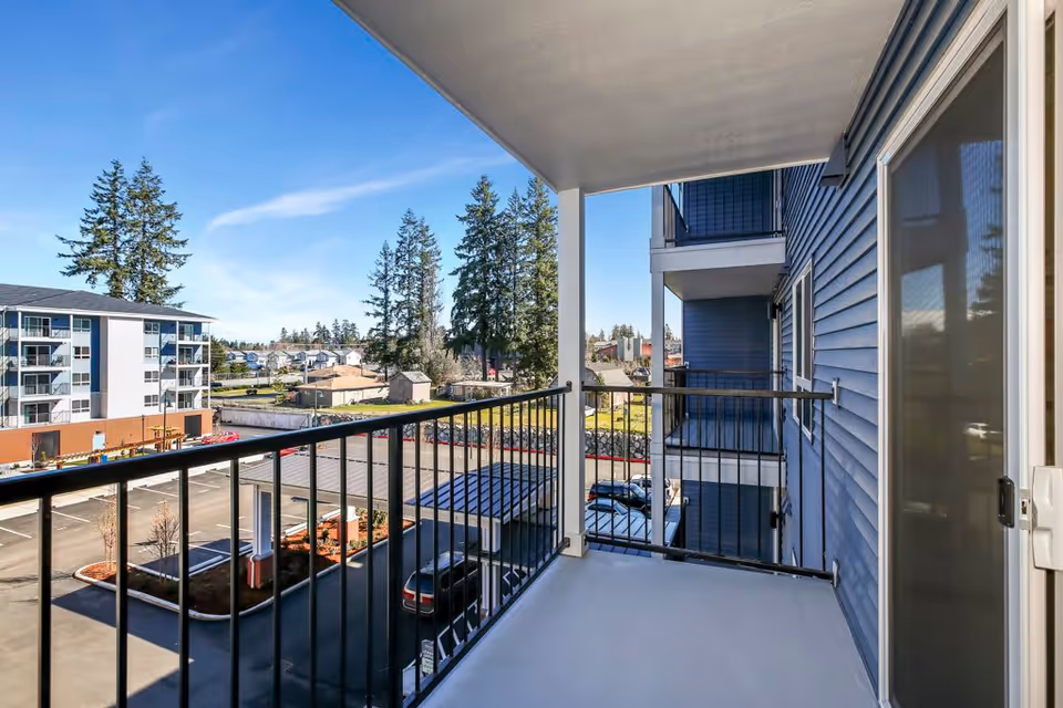 View from a covered balcony with black metal railing overlooking a parking lot, nearby buildings, and trees under a clear blue sky.