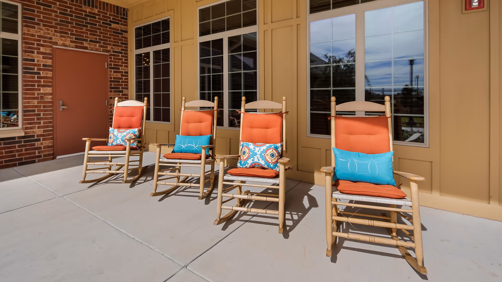Four wooden rocking chairs with orange cushions and colorful throw pillows arranged on a sunlit patio outside a building.