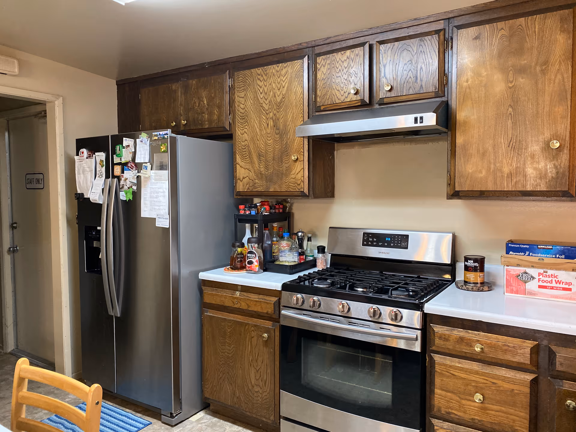 A kitchen with wooden cabinets, a stainless steel refrigerator covered with magnets and papers, a stainless steel gas stove with an oven, and various kitchen items on the countertops including spices, syrup, and food wrap boxes.