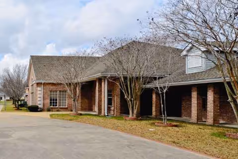 Brick senior living facility exterior with entrance, trees, and a parking area.