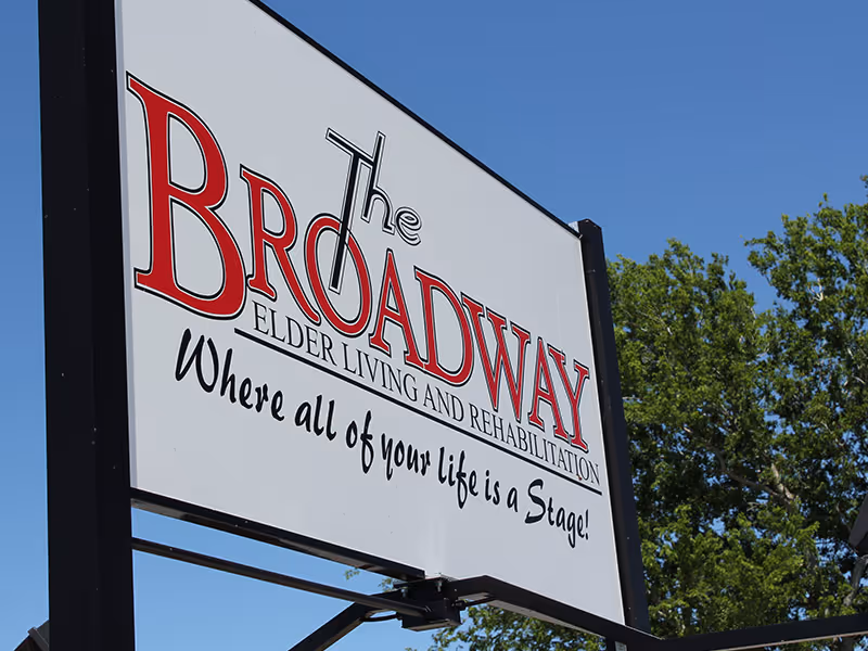 Outdoor sign for The Broadway Elder Living and Rehabilitation facility with the slogan 'Where all of your life is a Stage!' against a clear blue sky and some green trees in the background.