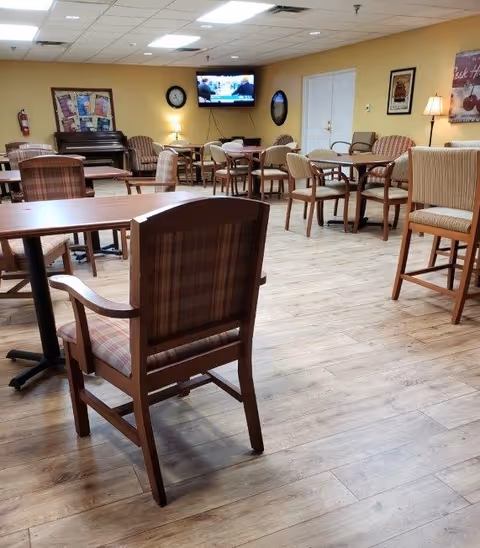 A senior living facility common area with multiple wooden tables and cushioned chairs arranged throughout the room. The floor is light wood, and the walls are painted a warm yellow. A piano is visible in the back left corner, along with a wall clock and a flat-screen TV mounted on the wall. There are framed pictures and a lamp on the right side of the room.
