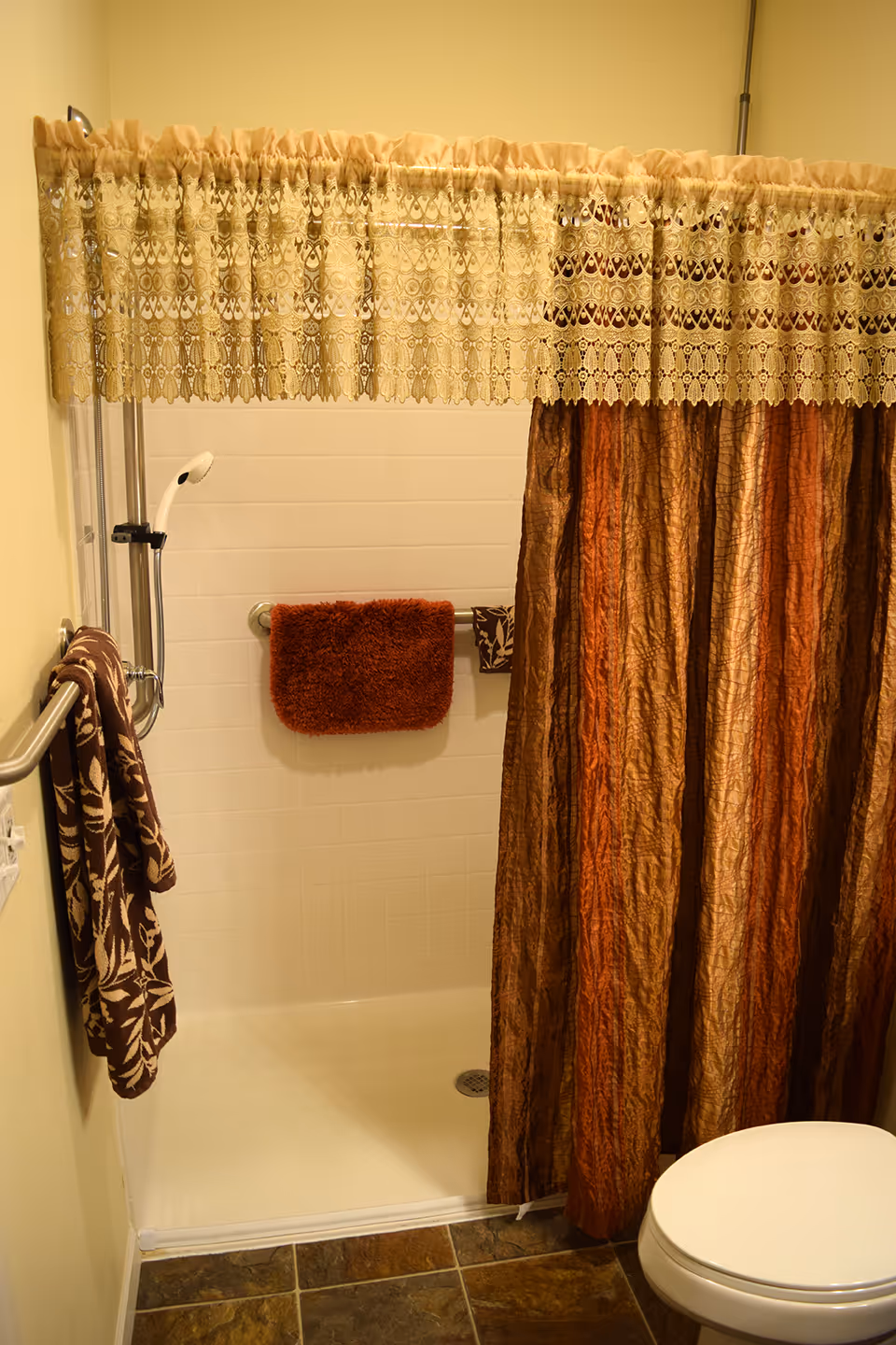 A bathroom shower area with a white tiled wall, a handheld showerhead, and a brown and orange striped shower curtain with a decorative lace valance above. There are two brown towels hanging on metal bars, one with a floral pattern and the other a solid rust color. The floor has dark brown tiles, and part of a white toilet is visible on the right side.