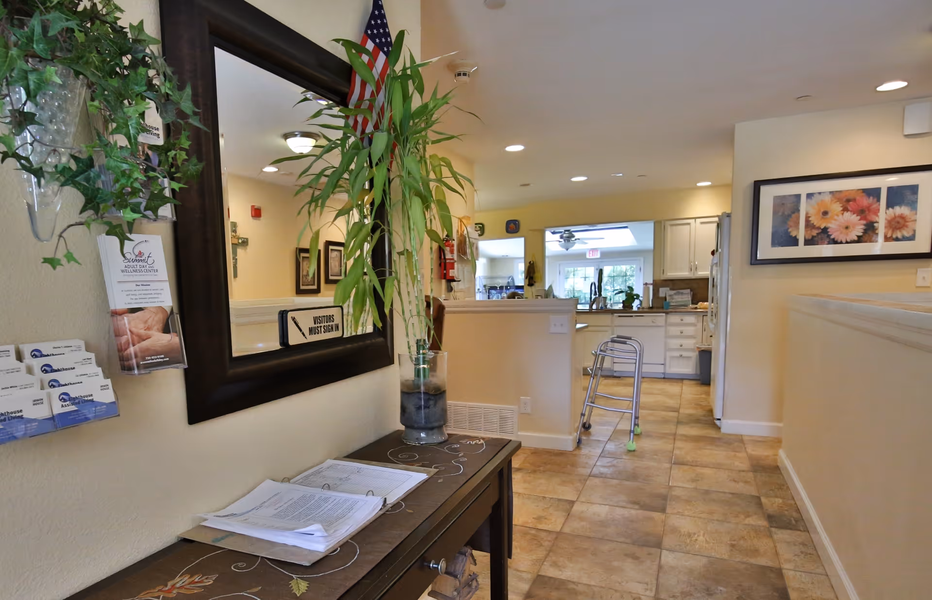 Interior view of a hallway in an assisted living facility showing a table with a plant, brochures, and a binder. A large mirror hangs above the table with a sign that says 'Visitors Must Sign In'. The hallway leads to a kitchen area with white cabinets and a walker is placed near the entrance to the kitchen. The walls are decorated with framed pictures and the floor is tiled.