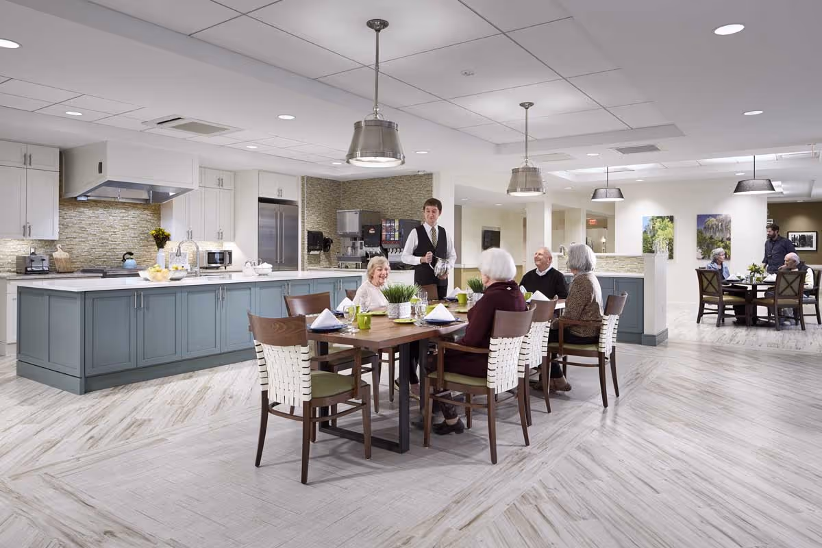 A modern dining area in a senior living facility with a large kitchen island in the background. Several elderly people are seated around a wooden dining table set with green cups and napkins, while a server in a vest and tie is pouring a drink. The room has light-colored flooring, pendant lights, and artwork on the walls.