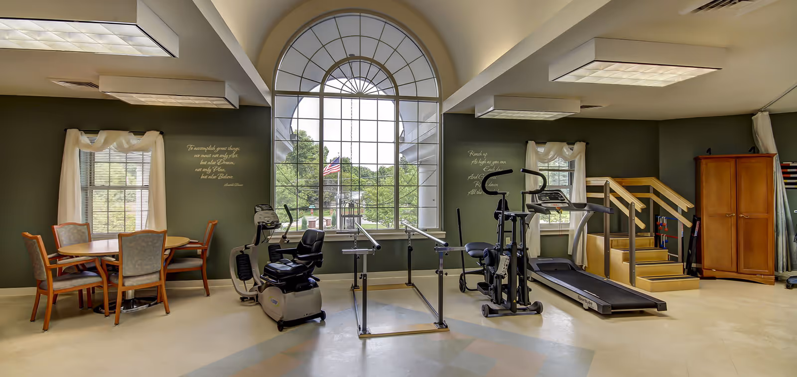 A well-lit rehabilitation or exercise room with large arched window showing greenery outside. The room contains exercise equipment including a recumbent bike, parallel bars for walking practice, an elliptical machine, a treadmill, and a wooden staircase with handrails for physical therapy. There is also a round table with four chairs near a window with white curtains. Motivational quotes are displayed on the walls.