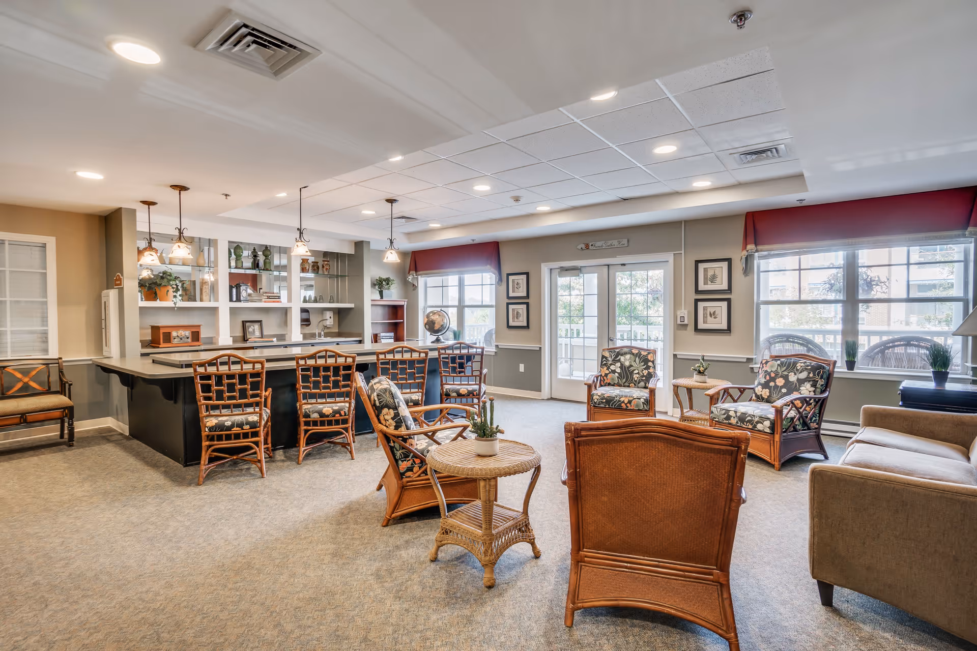A bright and spacious common area in an assisted living facility featuring a seating arrangement with wicker chairs and floral cushions around a small round wicker table. There is a beige sofa on the right side and a counter with four high chairs on the left. The room has large windows with red valances, framed artwork on the walls, and pendant lights hanging over the counter. A door with glass panels leads outside, allowing natural light to fill the room.