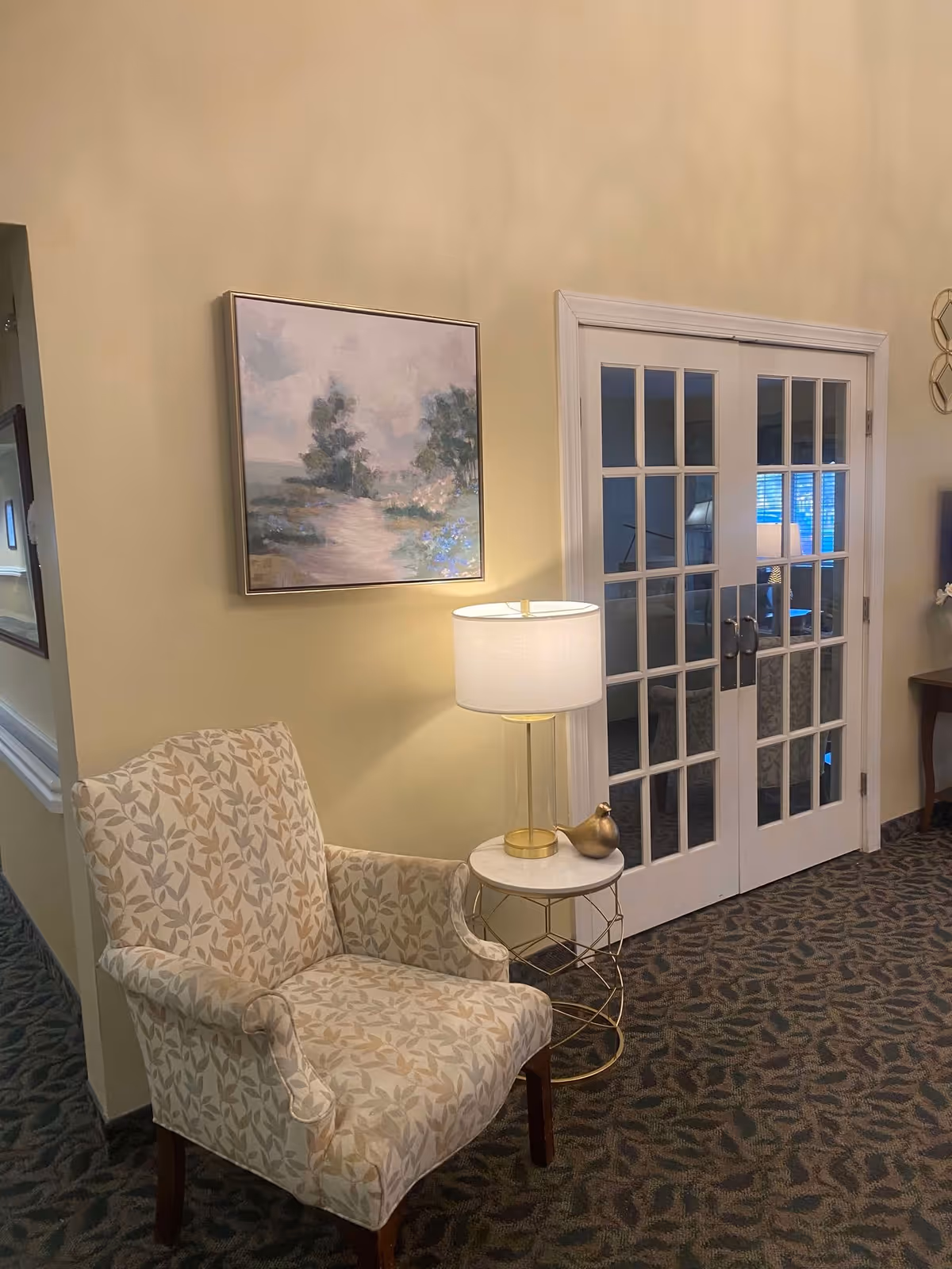 A cozy corner in a living area featuring a patterned armchair next to a small round side table with a white lamp and a decorative golden bird. Above the chair hangs a framed painting of a serene landscape with trees and a cloudy sky. In the background, there are white French doors with glass panes leading to another room.