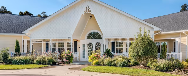 Front exterior view of a single-story senior living facility building with a peaked roof, white trim, and a covered entrance. The entrance has double glass doors with an arched window above, surrounded by well-maintained landscaping including bushes, flowers, and a small tree. The sky is clear and blue.