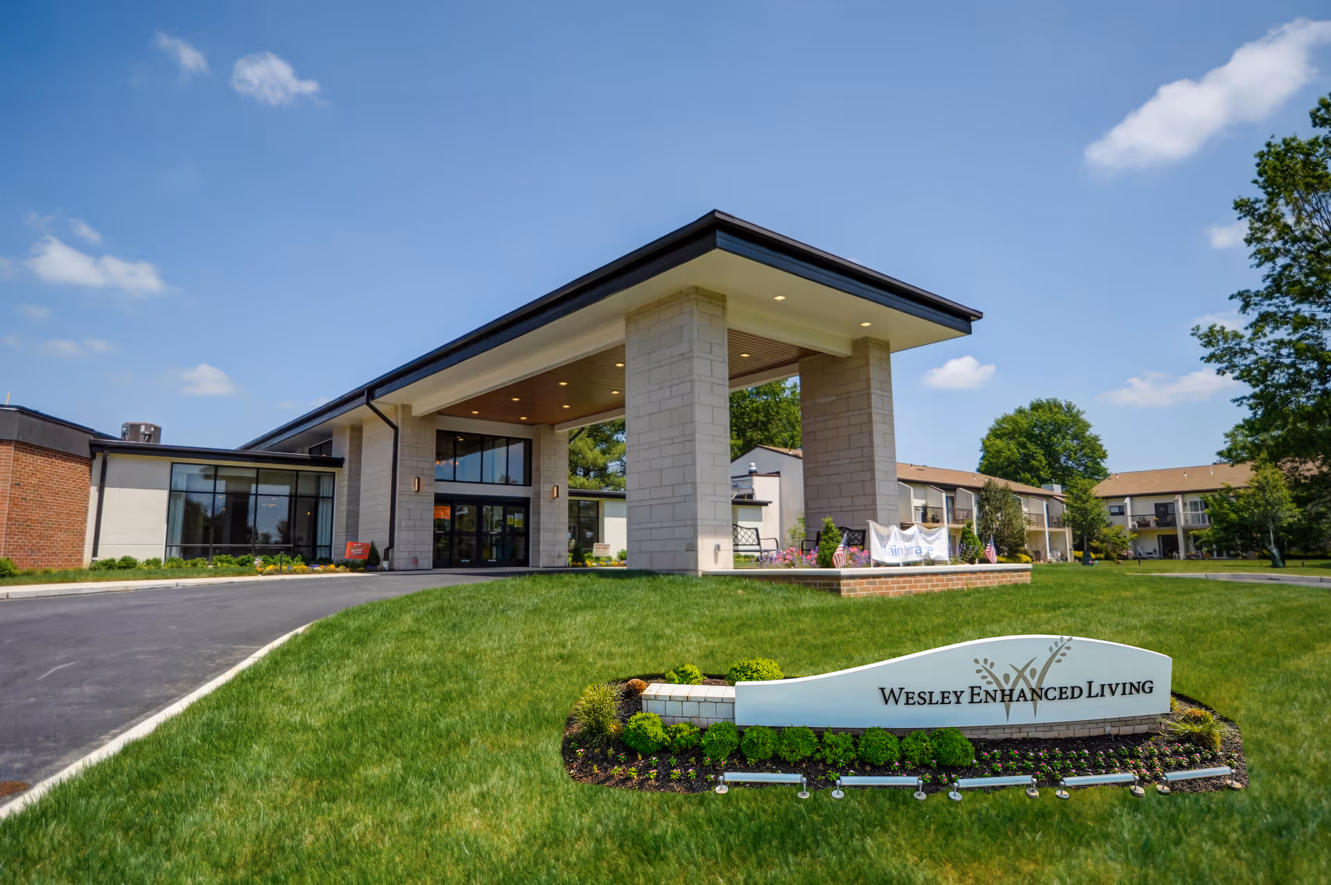 Exterior view of Wesley Enhanced Living Main Line facility on a sunny day, showing the main entrance with a covered driveway, well-maintained lawn, and a sign with the facility's name in front.