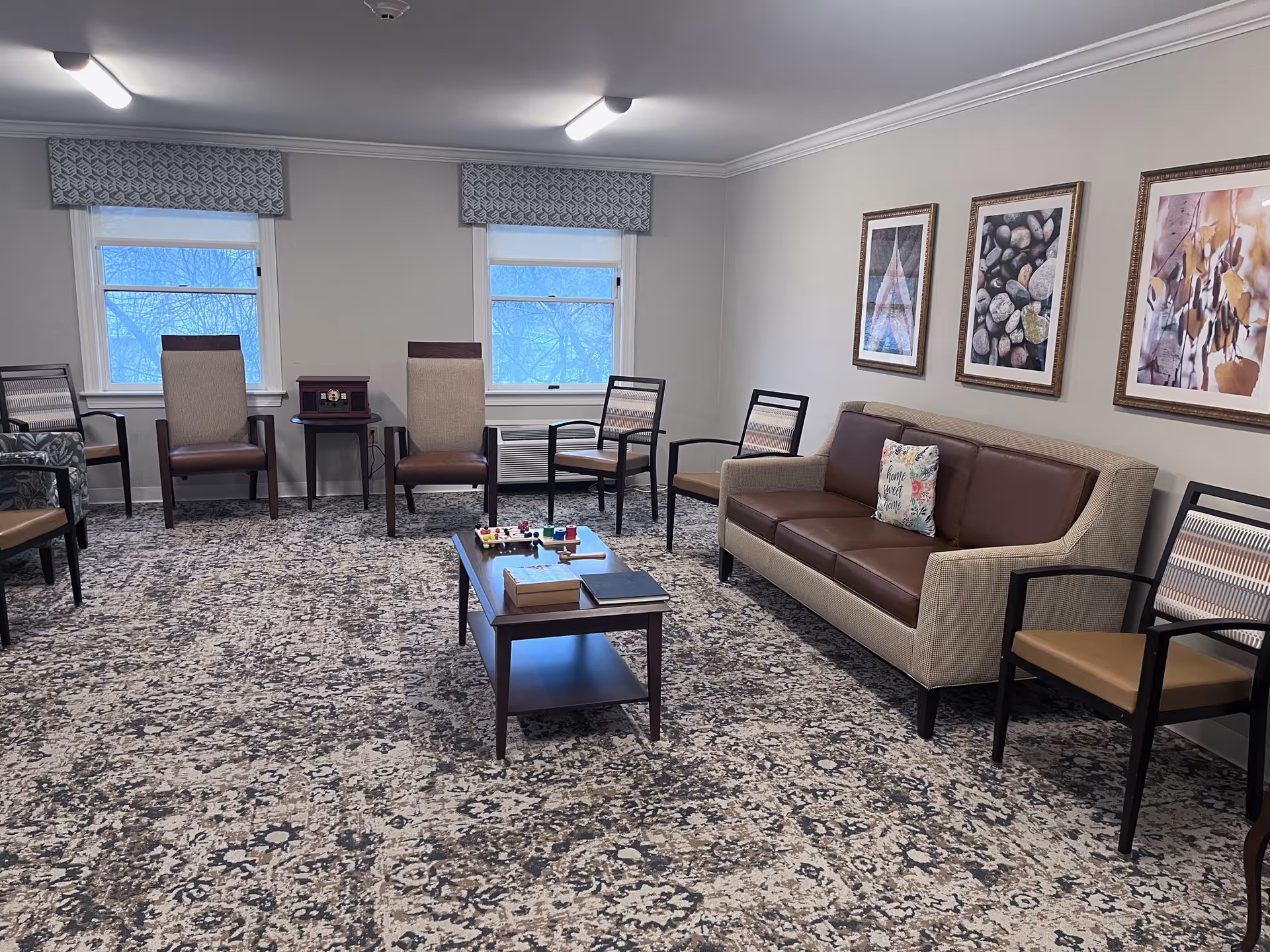 A senior living facility common room with a patterned carpet, a beige and brown sofa with a decorative pillow, several chairs arranged around the room, a wooden coffee table with books and games on it, and three framed pictures on the wall. Two windows with patterned valances let in natural light.