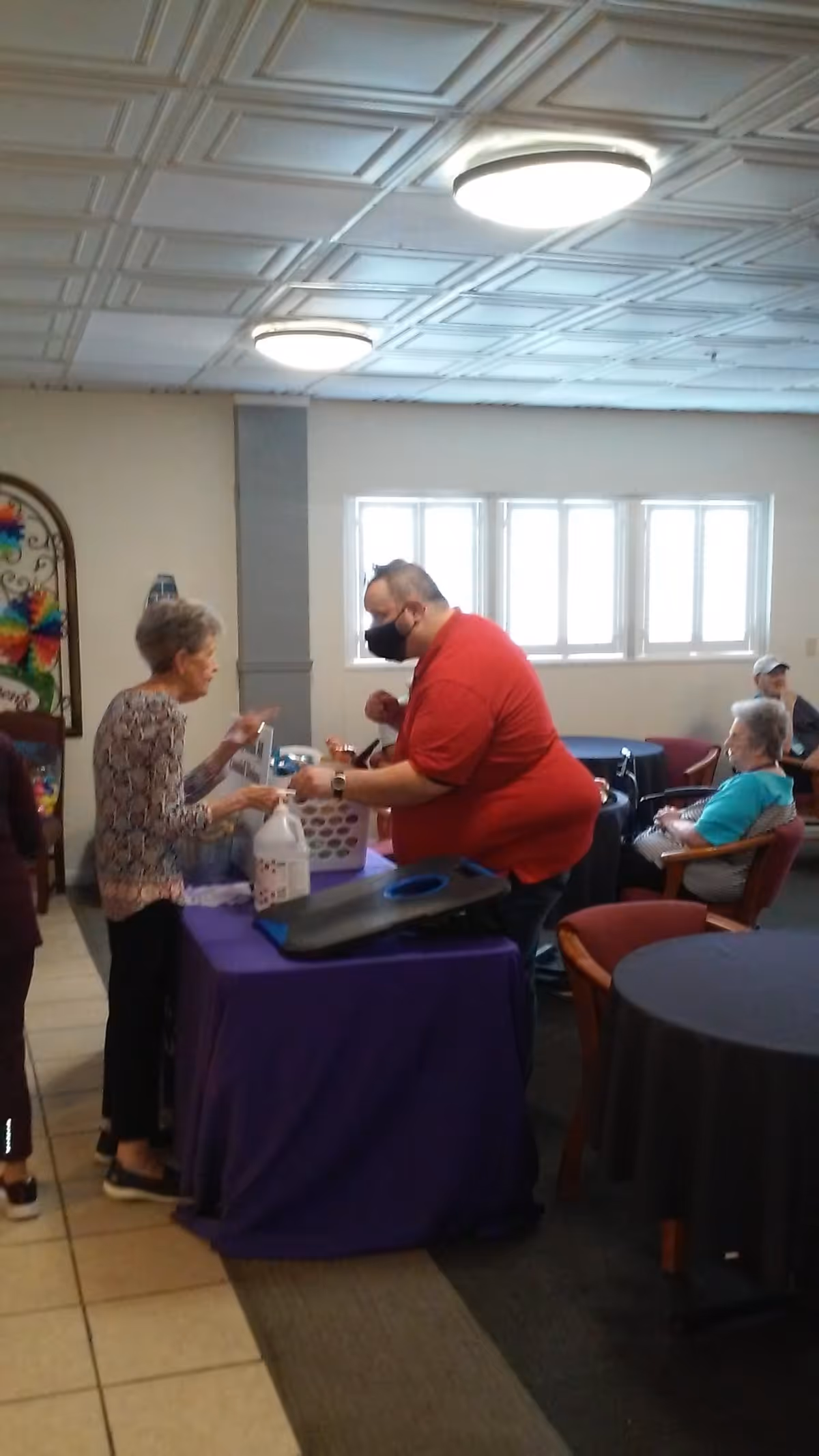 An indoor common area with several seniors interacting. A man wearing a red shirt and black face mask is standing behind a table covered with a purple cloth, engaging with an elderly woman. Other seniors are seated at tables in the background. The room has a tiled ceiling with round lights and windows letting in natural light.