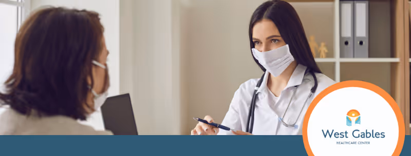 A healthcare professional wearing a white coat and face mask is speaking with a patient in an office setting. The healthcare worker holds a pen and appears to be explaining something. The background includes shelves with binders and decorative items.