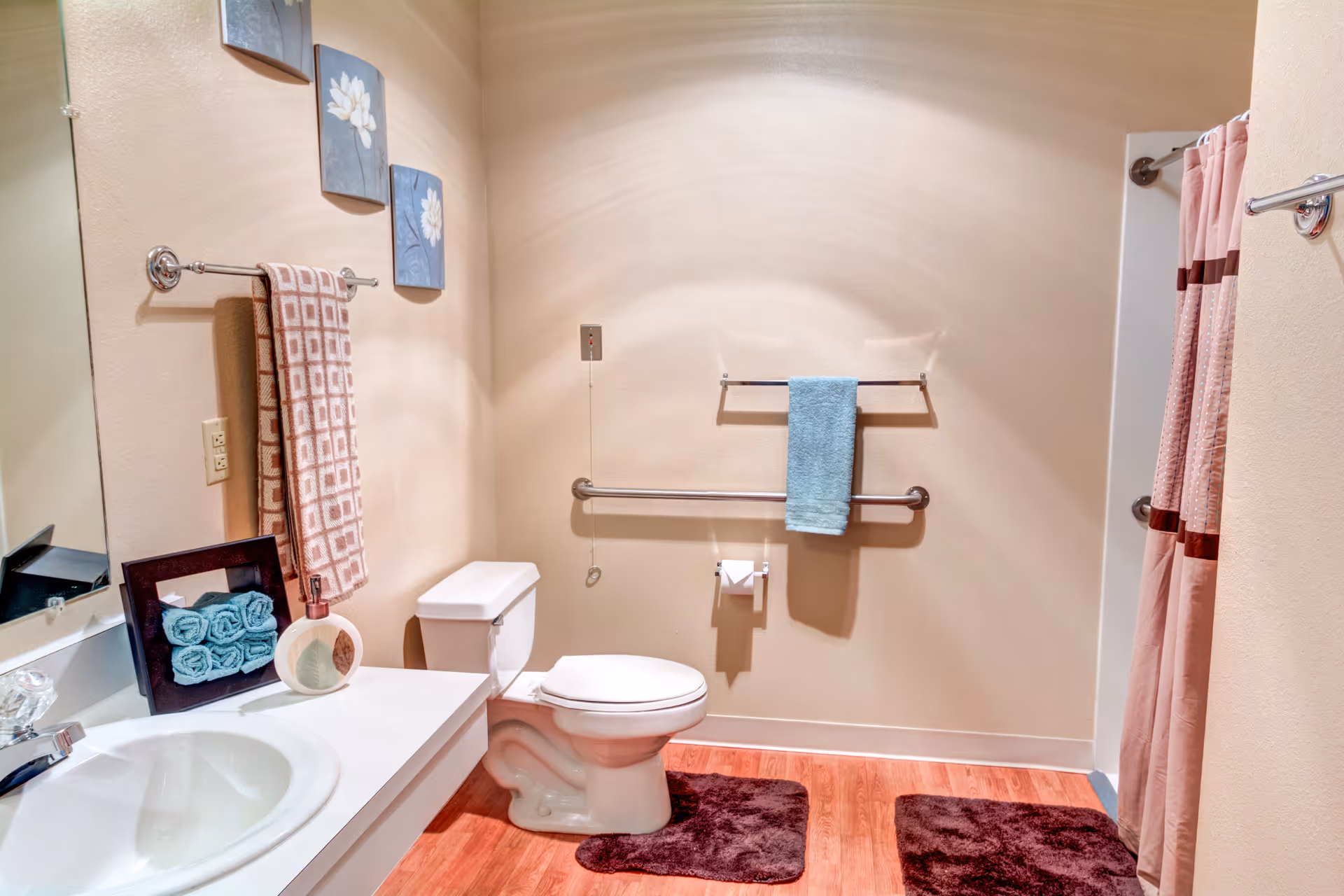 Well-lit accessible bathroom with a toilet, sink vanity, grab bars, towel racks and a shower with a pink curtain.