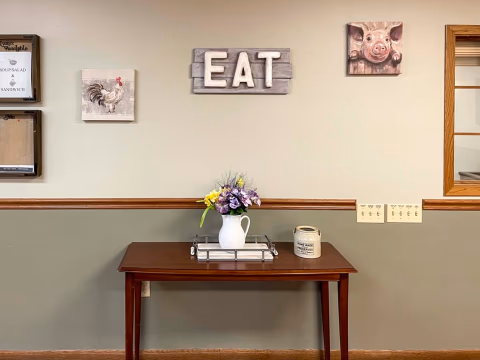 A wooden table against a two-tone wall with a white pitcher holding purple and yellow flowers and a small container labeled 'Home Made Preserves'. Above the table, there is a decorative sign with the word 'EAT' and two small paintings of a rooster and a pig. There is also a framed menu on the left and a window with wooden trim on the right.