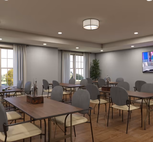 Communal dining/activity room with multiple wooden tables and gray upholstered chairs, windows, a wall-mounted TV, and a potted plant.