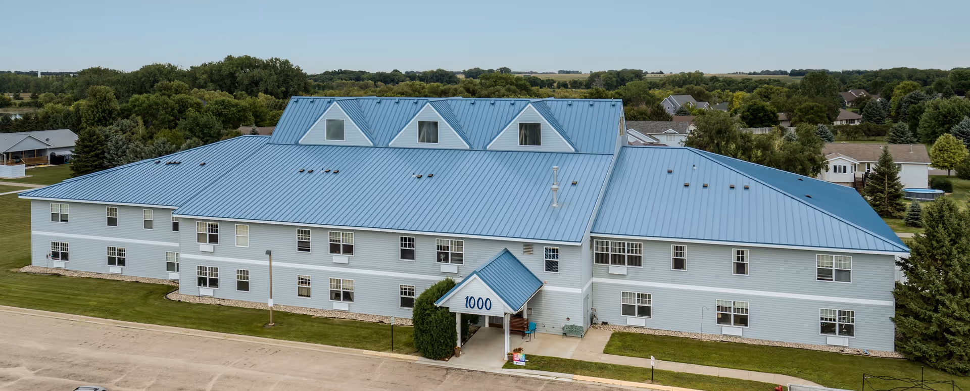 A large two-story building with a blue metal roof and light gray siding, surrounded by green lawns and trees. The building has multiple windows and a covered entrance with the number 1000 displayed. The area around the building includes a road and neighboring houses in the background under a clear sky.