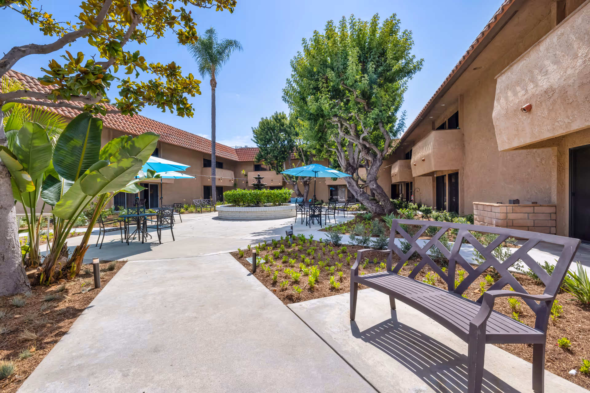 Outdoor courtyard area at Brookdale Garden Grove featuring a paved walkway, a metal bench, several tables with blue umbrellas, lush green trees and plants, and a central fountain surrounded by greenery. The courtyard is enclosed by a two-story building with a tiled roof under a clear blue sky.
