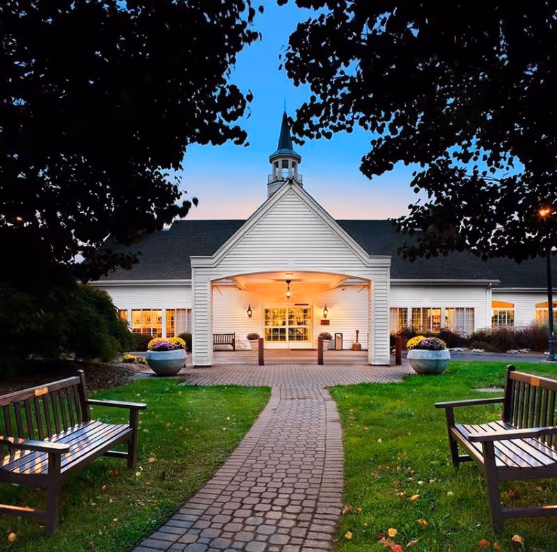 Front entrance of a white senior living building with a paved walkway, benches, and landscaped lawn at dusk.