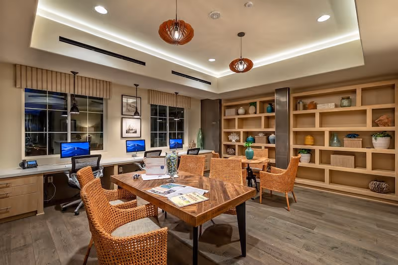 A well-lit interior room with a wooden table surrounded by wicker chairs, a built-in bookshelf with decorative items, and a row of desks with computers and office chairs along the wall under windows.