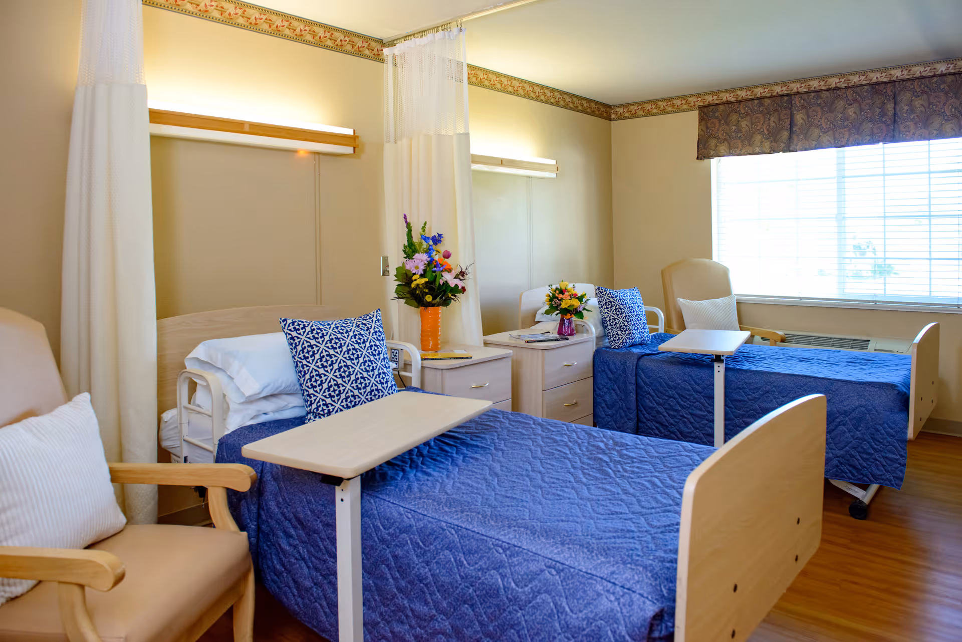 A bright and clean nursing facility room with two hospital beds covered in blue quilts, each with a blue patterned pillow. Between the beds are two nightstands with colorful flower arrangements. There are beige armchairs with white pillows next to each bed, a large window with blinds letting in natural light, and privacy curtains hanging from the ceiling.