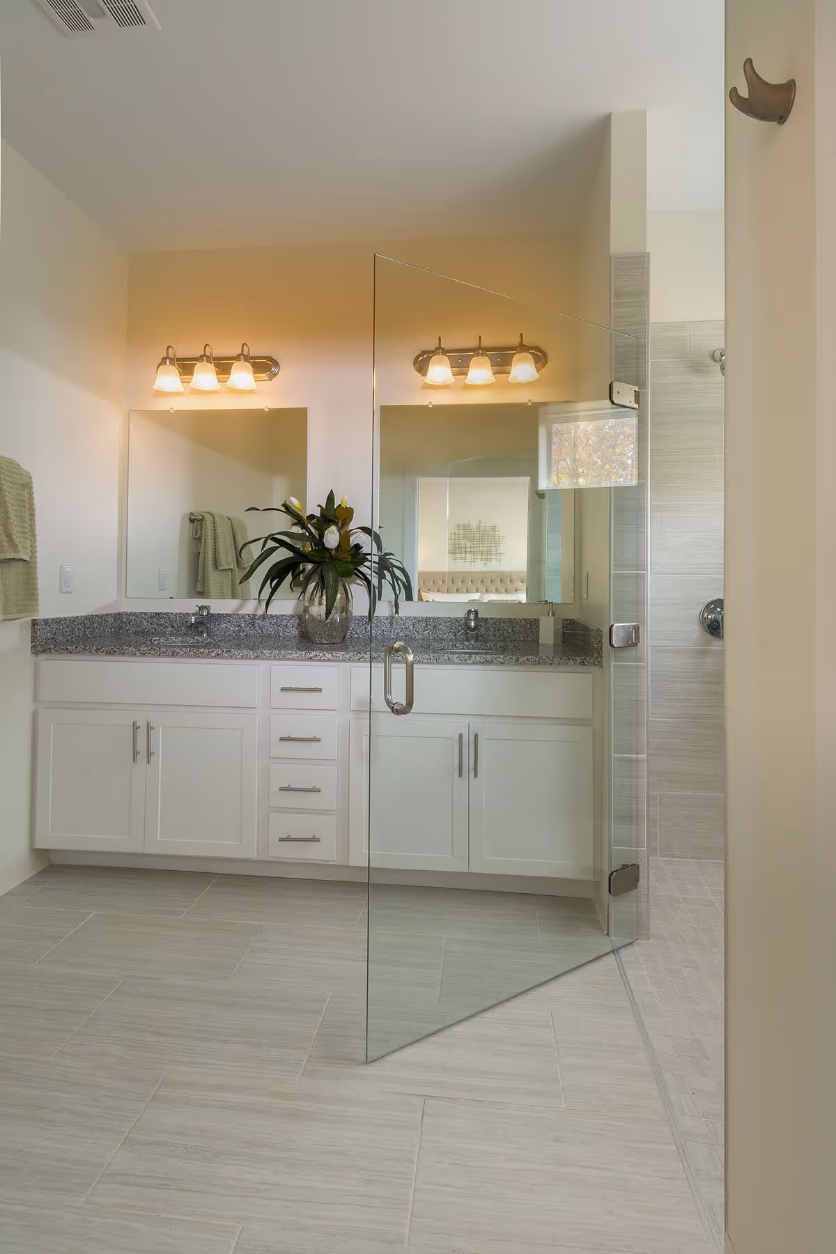 Modern bathroom with a double sink vanity featuring white cabinets and a granite countertop. Above the sinks are two mirrors with light fixtures. A glass door leads to a tiled shower area. A green plant in a vase is placed on the countertop.