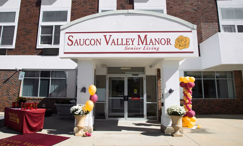 Front entrance of Saucon Valley Manor senior living building with a large sign, potted flowers, and balloon decorations.