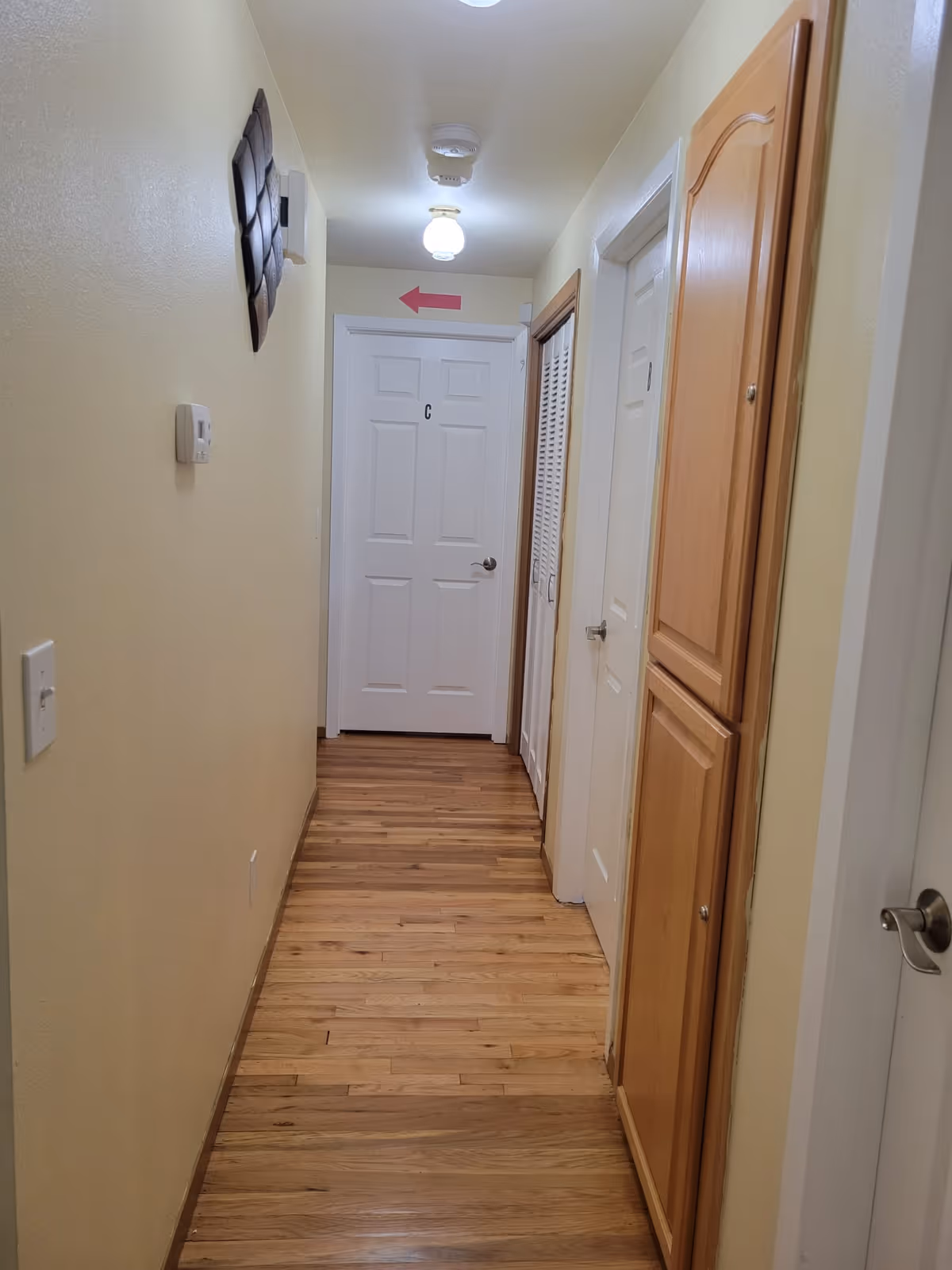 A narrow hallway with wooden flooring and cream-colored walls. There are several closed doors along the hallway, including a white door labeled 'C' at the end. A wooden cabinet is mounted on the right wall. The ceiling has two light fixtures and a smoke detector. A red arrow pointing left is visible above the door at the end of the hallway.