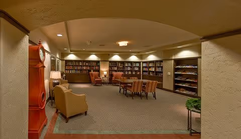 A cozy library or reading room with bookshelves lining the back wall filled with books. There are several comfortable chairs and a table with chairs in the center of the room. The room is softly lit with lamps and ceiling lights, and there is a large red clock on the left side near the entrance.