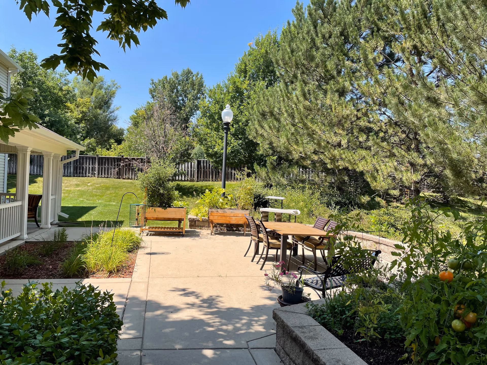 Outdoor patio area with a concrete walkway, wooden benches, a table with chairs, surrounded by greenery including trees, bushes, and plants. A white building porch is visible on the left side under a clear blue sky.