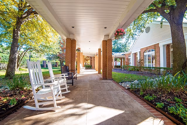 Covered outdoor walkway with brick columns and white ceiling, featuring white rocking chairs and benches along the path. The walkway is surrounded by green grass, trees, and landscaped garden beds next to a building with white siding and large windows.